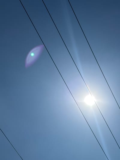 The image depicts a clear, light blue sky over San Juan Cuautlancingo, Mexico, viewed from a low angle. Multiple dark, thin utility lines or power cables traverse the frame diagonally from the upper left to the lower right. The sun is prominently featured in the lower-right quadrant, appearing as a bright, intense white light source with a starburst effect. A distinct lens flare is visible in the upper-left quadrant, manifesting as an elliptical, purple-tinted aura with a bright green central point. Additional faint linear light streaks emanate from the sun, extending across the sky. No people, specific subjects, actions, or interactions are present in the scene. The environment is outdoor, under daylight conditions.