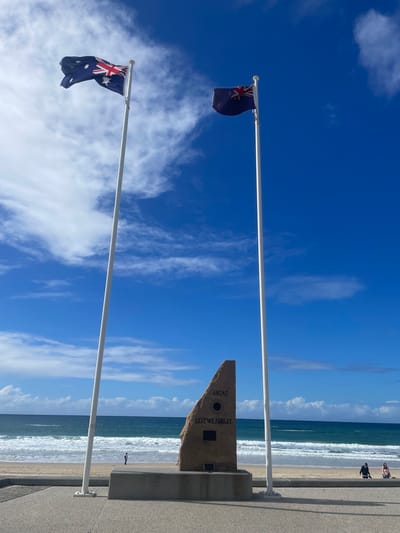 The image shows an Anzac memorial on the Gold Coast, Australia, with two flags flying. The memorial is a large, rough-hewn stone with the words "ANZAC" and "LEST WE FORGET" inscribed on it, along with two plaques. To the left of the memorial is a flagpole flying the Australian flag, and to the right is another flagpole flying the New Zealand flag. The flags are both visible in the image, with the Australian flag in the foreground and the New Zealand flag slightly further back. Behind the memorial and flags is a beach with waves rolling in from the ocean. A few people are visible on the beach and walking along the promenade. The sky is blue with some scattered clouds.