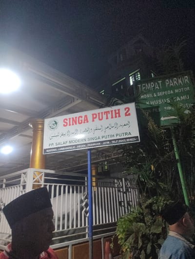 The image shows a sign for "Singa Putih 2 PP. Salafi Modern Singa Putih Putra" located at Sentol, Lumbangrejo, Prigen, Pasuruan, Jawa Timur, Indonesia. The sign is on a pole with a blue and white stripe and is surrounded by a white metal fence. There is a green sign for "Tempat Parkir Mobil & Sepeda Motor Khusus Tamu" to the right of the sign. The image was taken at night and shows a street light illuminating the sign. A person wearing a black cap is standing on the left, partially obscured by the sign. There is another person partially visible wearing a blue shirt on the right. There is a dark building behind the sign and a large amount of dark green foliage on the right.