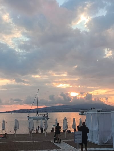This image depicts a serene beach scene at sunset. The sky is filled with dramatic clouds, illuminated by the warm hues of the setting sun, with patches of bright light breaking through. In the foreground, a sandy beach is lined with closed, white umbrellas, suggesting the end of the day or a break in activity. A few people are visible on the beach, some relaxing, others walking. One person is pushing a stroller, while another stands near a signpost.
The water is calm, reflecting the colors of the sky. A sailboat with its mast prominently in view is anchored or moored in the bay, with other boats visible in the distance. The shoreline is backed by a gently sloping hillside with some structures and trees, indicating a coastal town or resort area. The overall mood is peaceful and tranquil, capturing the beauty of a summer evening by the sea. The time of day is clearly sunset, with the soft, diffused light and warm color palette. The weather appears to be clear with scattered clouds.
On the signpost in the foreground, there is some visible text, though it is too blurry to be fully deciphered. It appears to be a list of prices or services, likely related to the beach amenities. The location is identified as Novoshakhtinsk, Russia.