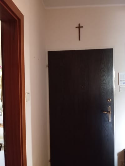A dark brown wooden door stands in the center of the frame, with a brass handle and lock. A small peephole is visible on the door. Above the door, a simple wooden cross is affixed to the white wall. To the left of the door, a white light switch is mounted on the wall. The wall extends to the left, revealing a portion of a wooden door frame and a darker wooden door. The scene appears to be an indoor hallway or entranceway.