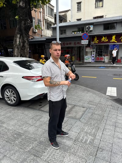 A young man stands on a city sidewalk in Liwan District, China, holding a clear plastic bottle of water. He is wearing a white short-sleeved shirt with a dark geometric pattern, black pants, and dark sneakers with red and white stripes. Sunglasses are perched on his head. He has tattoos on his left arm. The sidewalk is made of grey granite tiles and features tactile paving for the visually impaired. A white sedan is parked on the street behind him. Across the street, a row of shops with signs in Chinese characters is visible, one of which displays the words "汤粉" (tang fen), indicating noodle soup. A scooter rider wearing a yellow helmet is also present in the background. Buildings with windows and air conditioning units are visible beyond the shops. The sky is overcast.