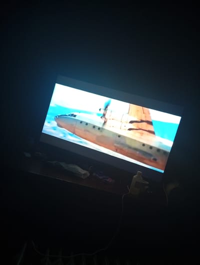 The image shows a television screen displaying an airplane in flight. The airplane is a propeller-driven aircraft, with its nose pointing towards the left and its engines visible on the wings. The background is a bright blue sky. The television is placed on a dark wooden surface, possibly a desk or table, which is littered with various objects. These objects include what appears to be a bottle of liquid with a red label, a white electrical cord, and some crumpled white paper. The lighting in the room is dim, with the light from the television screen illuminating the immediate surroundings. The overall scene is captured from a slightly elevated angle, looking down at the television and the objects on the surface below. No people are visible in the image. The location is identified as Suklieia, Moldova, based on the provided context.