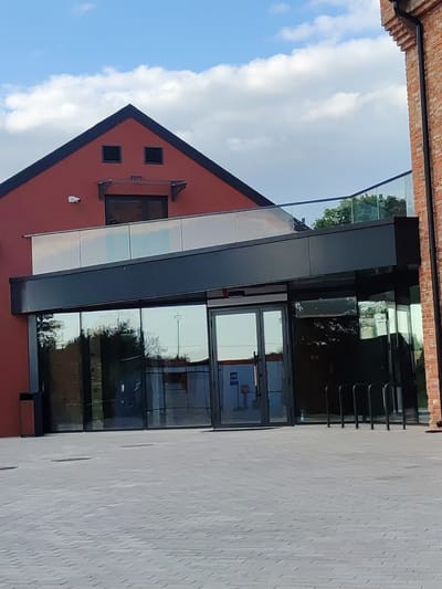 The image depicts the exterior of a modern building with a red facade and a dark, glass-fronted entrance. The building features a sloped roof with two square vents that resemble eyes, and a triangular gable. A security camera is visible on the red wall. A glass balcony railing runs along the upper level. To the right, a portion of a red brick building is visible. The foreground shows a paved area with a bike rack. The scene is set in Iisaku alevik, Estonia, under a blue sky with scattered clouds. No people are present in the image.