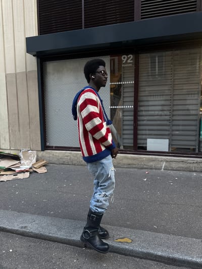 A person stands in front of a building in Saint-Ouen-sur-Seine, France. The individual, with dark skin and afro hair, wears glasses, a red-and-white striped sweater with blue trim, ripped light-wash jeans, and black boots. The building features a dark window frame and a metal roller shutter. "92 DUHESME" is printed on the glass. Debris, primarily cardboard, lies on the ground to the right of the person. The setting appears to be an urban street.