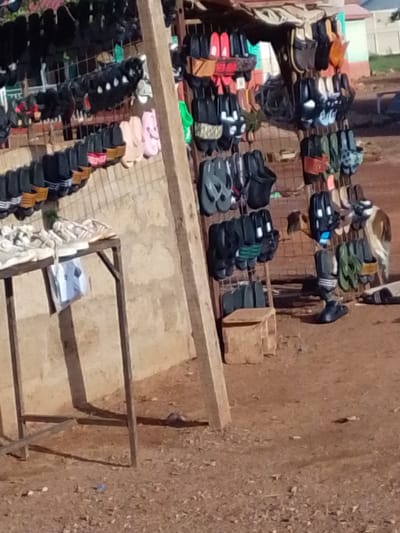 The image captures an outdoor shoe stall or market display in Dungu, Ghana, during what appears to be a sunny day. The primary subjects are a large assortment of sandals, slippers, and casual shoes, predominantly made of rubber or plastic, displayed on metal grids and racks.

The footwear is arranged in multiple rows and columns on two main vertical structures. Colors vary widely, including black, grey, red, pink, brown, and green, with some items featuring patterned straps or soles. Many items resemble popular brands of slides or flip-flops. On the left, a metal-framed table holds several white or light-colored shoes and what might be other goods, with a plastic bag hanging from its side. A sturdy wooden post supports the central display.

The ground is unpaved, consisting of dry, reddish-brown dirt or earth with scattered small debris. In the mid-ground on the right side, a light brown and white dog is visible near the base of the shoe displays. A small, light-colored wooden stool or box sits on the ground beneath some of the hanging shoes. The background includes a light-colored wall and glimpses of distant structures, indicating a semi-urban or rural setting. No specific text or branding is clearly legible on any of the items or structures. The scene depicts a typical vibrant, open-air retail setting.