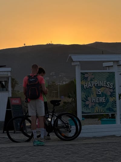 The image depicts a beach scene in Castelldefels, Spain, during sunset. The sky is a vibrant orange, casting a warm glow over the landscape. In the foreground, two individuals are standing next to bicycles on a sandy surface, which appears to be a boardwalk. One person, wearing a pink t-shirt and light-colored shorts, has a backpack. The other person is wearing a green t-shirt. To the left, a Coca-Cola sign is visible on a blackboard stand. To the right, a white structure with a sign displaying the text "HAPPINESS IS HERE" surrounded by tropical foliage is present. In the background, hills with scattered structures are silhouetted against the sky. The overall atmosphere suggests a relaxed, end-of-day setting.