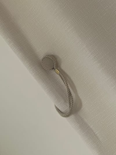 The image displays a detailed view of a light beige, textured fabric, consistent with a curtain or drapery. A light grey, braided cord magnetic tie-back is affixed to this fabric. The visible end of the tie-back features a circular, tightly coiled fabric disc, connected to the braided cord via a short, brass-colored metallic sleeve. The cord forms a curved loop, indicating its function in holding the fabric. A plain, lighter beige surface forms a diagonal background element. No individuals, actions, or additional environmental objects are present within the frame. This scene is located in Colombes, France.