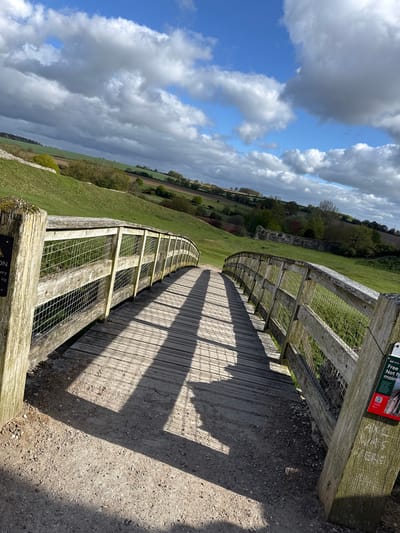 This image depicts a scenic outdoor view of a curved wooden boardwalk and adjacent gravel path, situated on a hillside or ridge. The boardwalk features sturdy wooden railings with wire mesh infill, casting long, distinct shadows onto the path and its own wooden planks due to bright sunlight.
To the left of the walkway, a vast expanse of green grass covers undulating hills, leading to distant fields and trees. In the midground, what appear to be ancient stone wall ruins are visible amidst the greenery. The sky above is a dynamic mix of clear blue patches and large, white and grey cumulus clouds, suggesting a partially sunny day, likely morning or late afternoon given the angle of the shadows.
No individuals are present, lending a peaceful and expansive feel to the scene. There are two visible signs attached to the wooden structure. One in the upper right is predominantly red and green, displaying a QR code and fragmented text including "Free", "Not from", and "mainte" (likely "maintenance"), possibly associated with a historical site or park. Another partially obscured sign in the bottom left shows text fragments like "...ON", "...ery", and "...icy", indicating a warning, potentially about "CAUTION" and "slippery" or "icy" conditions. The overall setting suggests a maintained path for exploration of a historical or natural landscape in the King's Lynn and West Norfolk area, United Kingdom.