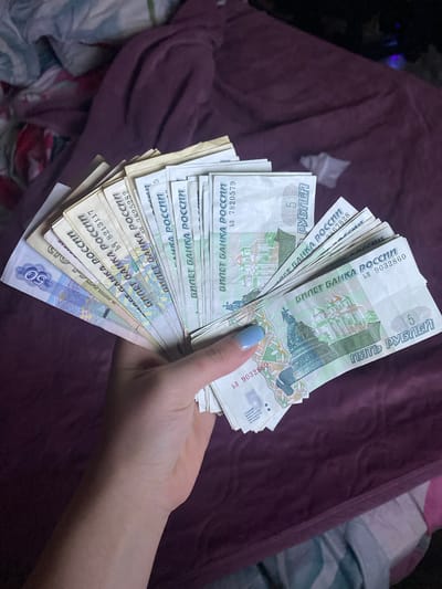 A close-up, dimly lit image shows a person's hand holding a fan of Russian banknotes against a dark purple, possibly velvet or plush, fabric background. The hand displays a thumb with light blue nail polish, gripping the stack of currency.

The majority of the visible banknotes are 5-ruble notes, identifiable by the text "ПЯТЬ РУБЛЕЙ" (Five Rubles) and the numeral "5". Prominent text "БИЛЕТ БАНКА РОССИИ" (Bank of Russia Note) and "РОССИИ" (Russia) are visible on multiple notes. Several serial numbers can be seen, including "ЫП 9032860", "b3 7820579", "БТ 2467828", "БТ 2825282", and "ЫП 8213117". One note at the bottom of the stack is clearly a 50-ruble note, marked with the numeral "50". Part of a security warning, "ПОДДЕЛКА БИЛЕТОВ БАНКА РОСС ПО ЗАКОНУ ПРЕСЛЕДУЕТСЯ" (Counterfeiting Bank of Russia notes is prosecuted by law), is also faintly visible on one note.

The setting appears to be indoors, likely a home, given the casual nature of the background fabric. The dim lighting suggests it could be evening or night. The currency explicitly indicates the location as Russia, aligning with the provided context of Verkhnevesyoloye, Russia. The activity is simply holding and fanning out money, possibly counting it or preparing for a transaction.
