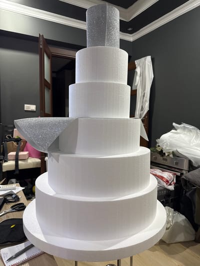 The image shows a six-tier foam cake display in what appears to be an indoor setting. The cake is primarily white, with the top and a mid-tier decorated with silver glitter. The cake is on a metal stand. In the background, a wooden door is visible, a dark gray wall, and various objects including a white garment hanging on a rack, plastic bags, a metal appliance, and a desk with a laptop, scissors, and a notebook. The flooring is light wood. There are no people in the image. The lighting suggests it is daytime. The location is identified as Gölbaşı, Turkey.