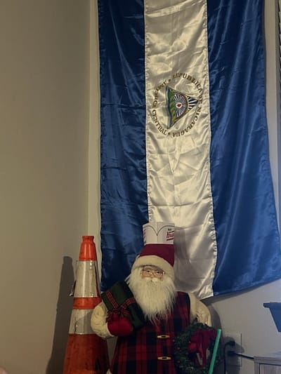 An indoor scene in Gainesville, United States of America, features a Nicaraguan flag hanging vertically on a light-colored wall. The flag consists of two horizontal blue stripes flanking a central white stripe, which prominently displays the national coat of arms. To the left of the flag, a plain light-colored wall is visible.

In the lower right portion of the image, a large decorative Santa Claus figure is positioned. The figure has a white beard, spectacles, a red Santa hat, and a white Krispy Kreme paper hat placed atop the Santa hat. It wears a red and black plaid vest and holds a green gift wrapped with a red ribbon in its left arm. A green wreath with red bows is visible near the figure's right arm. To the left of the Santa figure, an orange and white traffic cone, exhibiting signs of wear and dirt, leans against the wall. A white electrical outlet with a black power cord plugged into the upper receptacle is present on the wall to the right of the Santa figure. A green plastic object, possibly a straw or cup, is partially visible near the Santa's right arm.