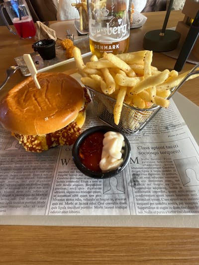 A top-down shot of a meal arrangement, likely in a restaurant, presenting a burger, french fries, and a drink. The burger, held together with a wooden skewer, sits alongside a wire basket overflowing with golden-brown french fries. A small black dish contains two sauces, ketchup and mayonnaise. The drink in a glass bottle, featuring the brand "Carlsberg" with "Copenhagen Denmark" on the label, is partially visible near the top left. The food is placed on a sheet of newspaper-print paper, which serves as a placemat. A red sauce container is also visible, partially obscured. The setting implies an indoor establishment, although no details reveal the location as Pirdop, Bulgaria, so that cannot be confirmed. No people are visible in the scene.