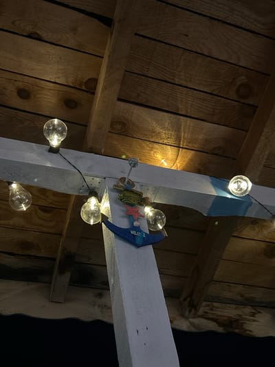 The image depicts a close-up, upward-facing view of a rustic wooden ceiling or roof structure, possibly an outdoor patio or covered area. Several clear, incandescent light bulbs are strung together by a black wire, illuminating the scene. The wooden beams have a rough-hewn texture, with visible grain and knots. A white beam, painted with a blue stripe, runs horizontally across the frame. Hanging from this white beam is a small, decorative anchor with a red starfish and a sign that reads "WELCOME." The lighting suggests it is nighttime or a dimly lit area, with the light bulbs casting a warm glow. The context provided, "in the city of Salmanovo, Bulgaria," indicates the geographical location of this scene. No people or specific activities are visible.