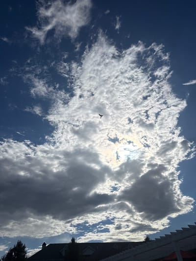 A wide-angle, upward-facing shot shows a dramatic sky over Allen Township, USA. Dark, heavy clouds dominate the lower half of the frame, contrasting with bright white, wispy clouds higher up. A small airplane is silhouetted against a bright patch of sunlight in the upper middle of the image. The sun's rays are visible, creating a lens flare effect. At the bottom of the frame, the roof of a house, trees, and a white pergola are visible.