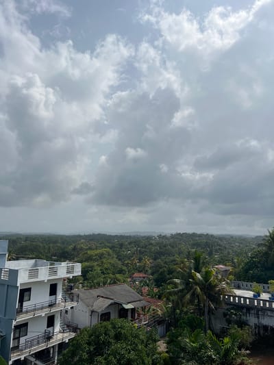 The image shows a view of Midigama, Sri Lanka, dominated by lush green foliage and a cloudy sky. In the foreground, a modern, multi-story building with white and blue facades stands to the left, featuring balconies with black railings and windows. Adjacent to it is a more traditional house with a grey tiled roof. Further into the scene, a series of buildings are nestled within the dense tropical vegetation. A prominent palm tree stands near the center right, with a wall topped with decorative white crenellations and potted plants in the background. The horizon is a hazy expanse of rolling hills covered in trees, suggesting a rural or semi-rural setting within Midigama. The sky is filled with dramatic, grey and white cumulus clouds, indicating potential for rain or an overcast day. No people or animals are visible in the frame.