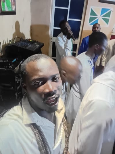 In the city of Ajibo, Nigeria, the image captures a group of men indoors, possibly at an event or gathering. In the foreground, a man with dark skin and short hair is looking directly at the camera with his mouth open. He is wearing a light-colored collared shirt and a necklace made of what appears to be beads. Behind him, several other men are visible. One man in a white shirt is holding a microphone, suggesting he might be speaking or performing. Another man in a blue garment stands further back. In the background, a sound mixing board is present, indicating potential musical or audio equipment setup. A framed picture hangs on the wall to the left, and a decorative geometric design adorns the wall on the right. The environment appears to be a dimly lit indoor space.