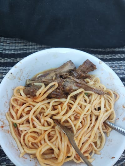 A white ceramic bowl containing a meal is depicted from a top-down perspective, situated in the city of Sidokepung, Indonesia. The bowl is filled with light orange-yellow, thick-cut noodles coated in a sauce, topped with several pieces of dark brown, braised bone-in meat. A small amount of dark brown broth or sauce is visible around the meat and noodles. A silver-colored fork is partially submerged in the noodles on the left side of the bowl, and a silver-colored spoon is partially visible on the right. The white rim of the bowl shows minor food residue. The bowl rests on a surface with a black and light grey horizontally striped or woven pattern. The background above the bowl is a dark, blurred, black fabric texture with faint specks. No individuals are present in the image.