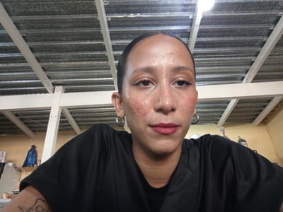 A medium-shot, low-angle selfie of a person with dark hair slicked back, wearing silver hoop earrings and a black short-sleeved shirt. The person has a slightly flushed complexion and is wearing makeup. The background consists of a corrugated metal roof supported by white wooden beams and a light fixture. The room's walls appear to be painted pale yellow. In the background, on the left, a small, blue statue can be seen on a shelf. The image was likely taken indoors in El Dátil, Venezuela.