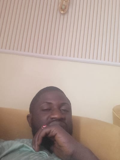 A low-angle, interior close-up shot depicts an adult male of dark complexion, identified as being in Babawa, Nigeria. The individual occupies the lower-center frame, partially obscured by his right hand, which is raised to his chin/mouth area. A ring with a prominent reddish stone is visible on his right ring finger. He wears a light green or teal-colored garment. His eyes appear closed or heavily lidded. Behind the subject, a mustard-yellow upholstered surface, likely a couch or armchair, is visible. The background consists of a plain, light yellow wall transitioning to a ceiling paneled with vertical, light-colored slats. A translucent, possibly amber-toned light fixture or decorative element with a white interior component is affixed to the paneled ceiling in the upper right quadrant.