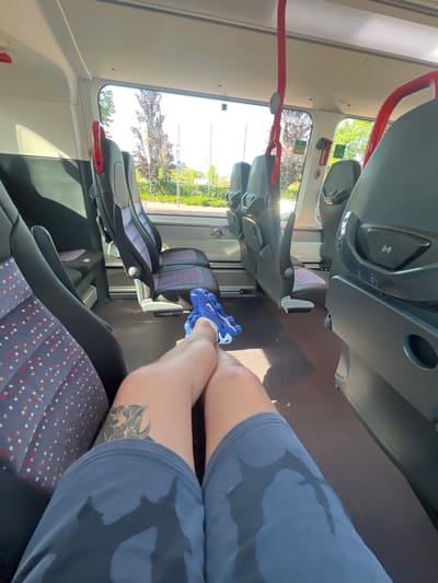 The image depicts an individual on a train. The individual is wearing blue shorts and a blue shoe, and they have a tattoo on their left calf. The train seats are gray and red. The window shows a view of the exterior, including trees and what appears to be a street. Based on the available information, it's not possible to confirm the specific location within Oud-Beijerland, Netherlands.