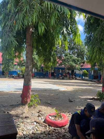 In Turumame, Indonesia, an outdoor scene unfolds in what appears to be a schoolyard. The foreground is dominated by the base of a tree with its trunk painted red and blue. Scattered around the tree are rocks, broken bricks, and a red-painted tire serving as a planter for a small plant. To the right, three young individuals wearing blue vests and dark caps are crouched down, seemingly engaged in an activity. One individual's cap displays a yellow emblem.

In the mid-ground, a paved area, possibly a basketball court, is visible. Beyond this, a blue and white building with red-tiled roofs lines the perimeter of the yard. Several students, dressed in uniforms, are seated on a low wall or steps in front of the building, some appearing to converse. Other students are standing or walking in the vicinity of the building.

The background features more trees, with lush green foliage, and a bright blue sky with scattered white clouds. The overall impression is a sunny day in a school environment with a moderate number of students present.