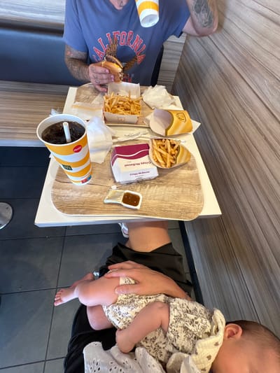 A group of people are dining at a McDonald's in Carini, Italy. The table is laden with food and drinks, including a burger, fries, and a cola. A baby is being held by one of the diners, its tiny feet visible. The scene appears to be casual and relaxed, with the group enjoying their meal. The time of day is not specified, but the lighting suggests it might be daytime. The interior of the McDonald's is visible, with wooden tables and tiled floors. The presence of McDonald's branding indicates the location.