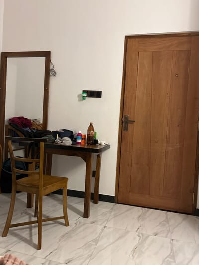 The image depicts an interior scene, possibly a bedroom or hallway, in Kadewatta, Sri Lanka. A large wooden door dominates the upper portion of the frame, featuring horizontal planks and a metal latch. To the right of the door, a floor is covered with marble-patterned tiles.
In the lower half of the image, a white wall is visible. Mounted on the wall are electrical outlets and a small green dispenser. In front of the wall, a wooden shelf unit holds several items, including bottles, bags, and what appear to be hats or caps. A wooden chair with an open back is placed beside the shelf unit, with its legs extending onto the tiled floor.
Further to the left, a wooden-framed mirror is leaning against the wall. The overall impression is of a functional, perhaps slightly cluttered, living space. The lighting suggests daytime, but no specific time or weather conditions can be inferred. There is no discernible text in the image.
