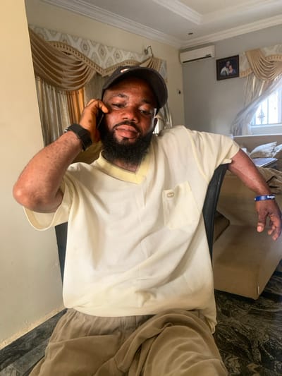 The scene depicts an indoor residential room located in Eika-Adagu New Layout, Nigeria. A single adult male, appearing to be 20-40 years old, with dark skin and a full beard, is seated and facing forward. He wears a black baseball cap, a light cream short-sleeved V-neck shirt with a left chest pocket featuring a small metallic emblem, and light brown trousers. A black digital watch is present on his left wrist, and a blue wristband with white text is on his right wrist. His left hand is raised, holding a dark-colored smartphone to his left ear, indicating he is engaged in a phone call. He looks directly towards the camera. The background consists of light-colored walls, white crown molding, and elaborate gold-toned curtains with valances. The flooring is dark, mottled, and appears to be polished tile. An air conditioning unit is mounted high on the right wall, above a window fitted with security bars. A framed picture hangs on the wall, and a light-colored sofa is partially visible on the right side of the frame. The room is illuminated by ambient light, suggesting a daytime setting.