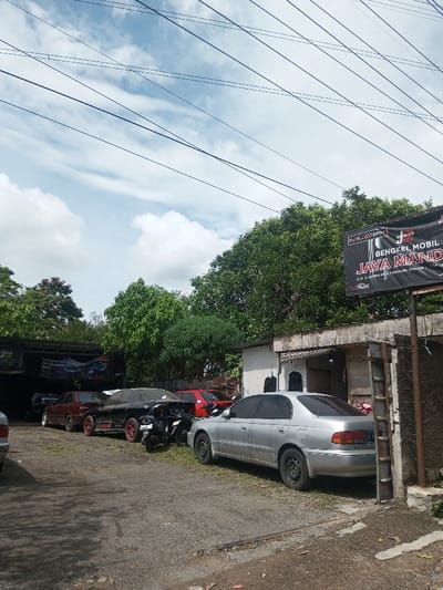 A row of cars and a motorcycle are parked in front of a building marked "Bengkel Mobil Jaya Mandiri." The vehicles are varied in color, with a silver sedan closest to the camera. Overhead, a network of power lines crisscrosses the sky. Lush green trees form a backdrop to the scene, contrasting with the partly cloudy sky above. The location appears to be a roadside automotive repair shop.
