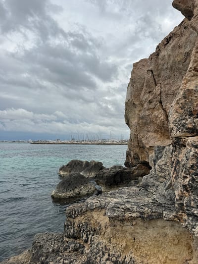 The image depicts a rocky outcrop overlooking the sea in Llucmajor, Spain. The perspective is from within a cave or under a rock overhang, framing the scene. The sky is overcast with grey clouds. In the distance, a town is visible along the shoreline, characterized by masts of boats suggesting a marina or port area. The sea appears calm with varying shades of blue and grey. The rocky formations in the foreground are textured and rugged, showing signs of erosion and weathering. There are no people visible.