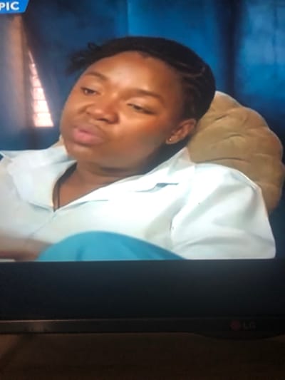 An adult female, characterized by a dark skin tone and short, dark braided hair, is depicted from the chest up in an indoor setting within the city of Masaka, Nigeria. She wears a light-colored, collared shirt, unbuttoned at the top, over a dark garment, with teal or light blue fabric visible below the shirt. A small, light-colored stud earring is present in her right earlobe. She is seated or reclining, resting her head against a light brown, padded object that serves as a headrest. Her head is tilted slightly down and to the left, with her eyes partially closed or looking downwards, and her lips are slightly pursed. The background is a dark blue wall or curtain, with a blurry, vertical lighter element visible on the left. The image itself is a screen capture, indicated by a partially visible "LG" logo on the bottom right bezel and a "PIC" logo in the upper left corner.