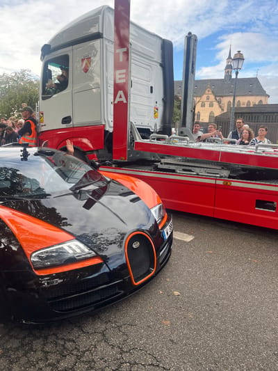 The image depicts an outdoor scene in Molsheim, France, with a prominent historic building featuring multiple spires and a brown roof visible in the background, characteristic of structures like the Église des Jésuites.

In the foreground, a two-tone black and orange Bugatti sports car is positioned on an asphalt road with visible cracks and sparse leaves. The vehicle's front section is black with distinct orange accents on the hood, front fenders, and around the iconic horseshoe grille. A French license plate displaying "W-388-GS" is visible on the front bumper. A small black camera or sensor device is mounted on the vehicle's roofline near the windshield. From the driver's side of the Bugatti, a human hand is extended upwards and outwards, performing a wave.

In the midground, to the right of the Bugatti, a white and red semi-truck, configured as a car transporter, is present. The truck's cabin is white, with a red lower section and chassis. An "ALSACE" crest is affixed to the driver's side door. A tall, red vertical beam or panel behind the cabin features the word "TEA" in white capital letters. The truck's red flatbed platform is visible, appearing partially elevated.

Several individuals are present. Inside the truck's cabin, a person, presumably the driver, is visible holding a smartphone in a photographic stance. A crowd of spectators is gathered on the far left, partially obscured, with one man clearly visible in an orange high-visibility vest and a baseball cap, facing towards the right. On the raised platform of the truck's flatbed on the right, a group of approximately five spectators is observing the scene. This group includes two younger males actively using smartphones to take photos or videos, and three adults (two females, one male) watching.

The sky above is partly cloudy with patches of blue. A black, ornate street lamp post stands in the midground on the right, in front of the historic building.