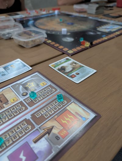 This image shows a tabletop board game in progress on a wooden surface. The components visible include: a circular game board with a Martian landscape and various tokens, cards with text and images, personal player boards with resource tracking, and translucent dice-shaped markers indicating progress along the resource tracks. Specifically, one card shows a location called "MOHOLE AREA". Some cards appear to be discarded or awaiting use. The player boards have different sections for different resources.
