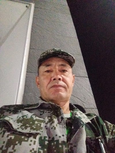 This is a selfie of an adult East Asian male, who appears to be middle-aged, wearing a camouflage cap and a camouflage jacket over a white t-shirt. He is looking directly at the camera. The background is a textured gray wall with a darker section on the right. To the left, there is a white vertical element. The lighting suggests it could be evening or indoors with artificial lighting. No text is visible in the image. The setting appears to be an urban or semi-urban environment, possibly a public space or a building interior. The man's expression is neutral. The provided location context indicates he is on the Northern Bypass Road 84 km in Kyrgyzstan.