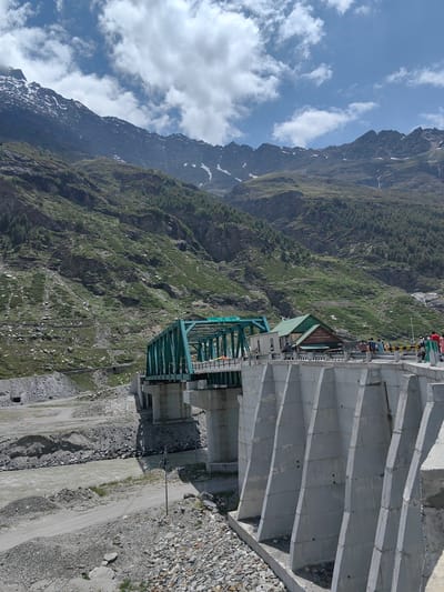 The image depicts a concrete dam structure on the right, with a green truss bridge spanning a river in the background, likely near the city of Burwa, India, given the context. The dam is constructed with large, trapezoidal concrete segments. Beyond the dam, the green truss bridge supports a road and a small building. Several people are visible on the bridge and near the building, appearing to be tourists or locals.
The environment is mountainous, with steep, green slopes covered in vegetation and scattered rocks, rising to jagged peaks in the distance. Some of the higher peaks show patches of snow. The sky is blue with scattered white clouds. A muddy river flows beneath the bridge. The ground in the foreground consists of gravel and rough terrain. The scene suggests a scenic, possibly tourist-oriented location within a mountainous region. The presence of the dam and bridge indicates infrastructure development in the area.