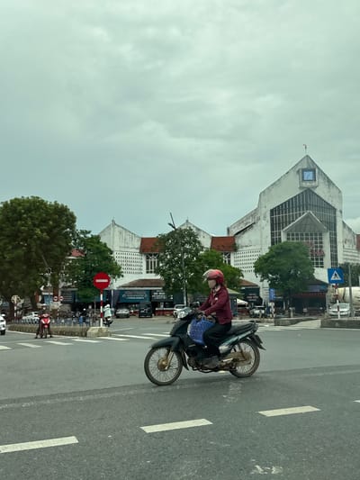 A medium-shot, eye-level, outdoor, daytime, vertical photograph captures a street scene in Nam Đông Hà Ward, Vietnam. The foreground features an asphalt road with white lane markings. A person wearing a red helmet and a maroon jacket rides a dark green motorcycle across the road, heading towards the right. The motorcycle has a blue plastic basket attached to the front. In the middle ground, a pedestrian crossing is visible with a red circular traffic sign indicating prohibition. Several other motorcycles and cars are present on the road and sidewalk. Behind the road, a large, multi-story white building with a distinctive architectural style stands out. The building has a triangular roof with a clock face and a flag on top. There are trees and other structures flanking the main building, suggesting a commercial or public area. The sky is overcast with visible clouds, indicating a dull weather condition.
