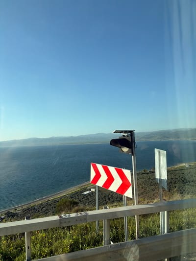 The image captures a bright, sunny daytime landscape, likely viewed from a roadside elevated vantage point overlooking a large body of water. The perspective is from an upward angle, showing a clear blue sky dominating the upper left portion of the frame.

In the mid-ground, a vast expanse of dark blue water stretches horizontally across the image, exhibiting subtle ripples on its surface. Along the water's edge, a narrow strip of land is visible, covered with dense green and brown vegetation, possibly including trees and shrubs, and hints of a distant road or path. Further across the water, a hazy outline of mountains or hills can be seen, fading into the light blue horizon.

The foreground, on the right side of the image, features robust roadside infrastructure. A sturdy metal guardrail system, composed of horizontal beams and vertical posts, extends across the frame. Green grass and scattered yellow wildflowers grow alongside the barrier. A black, cone-shaped traffic signal or light fixture is mounted on a metal pole, possibly equipped with a small solar panel on top, suggesting it's self-powered. Below it, a prominent white sign with three red chevron arrows pointing to the right indicates a turn or hazard. Another smaller, plain rectangular white sign is also visible.

No human activity or specific event is taking place, presenting a static, serene view. The bright daylight and clear sky indicate good weather. On one of the metal posts of the guardrail, numbers "156" and "560" are visible, possibly identifying markers for the infrastructure. Given the context, this scene likely depicts Lake Iznik in Turkey.