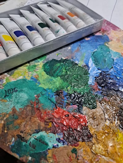 This media file features a close-up shot of a painter's palette laden with thick, textured oil paints in a vibrant array of colors, including blues, greens, reds, yellows, and browns. To the left of the palette, a box of paint tubes is visible, some of which have their labels showing. The text on the tubes includes "Boby", "Acrylic Color Green", "Boby Acrylic Color Marine Blue", and "Boby Acrylic Color Yellow". The overall impression is one of artistic creation in progress, with the rich colors and textures suggesting a dynamic and expressive painting. The scene appears to be an indoor setting, likely an artist's studio or workspace. There are no people visible in the frame, and the focus is entirely on the art supplies. The lighting suggests it could be daytime, but there are no explicit cues to determine the time of day or weather conditions. The location context of Popovo, Bulgaria, is provided but is not visually discernible from the image itself.
