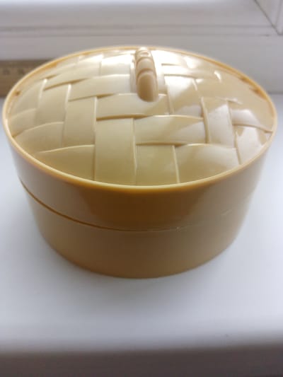 A light brown, plastic container with a woven-pattern lid sits on a white surface, likely a shelf or table. The container has a smooth, cylindrical body with a slightly wider rim. The lid is curved and features a criss-cross pattern made of raised strips, resembling a basket weave, with a small handle or knob on top. The background is blurred, with a light-colored wall or surface visible behind the container and a darker horizontal line, possibly the edge of a ruler or shelf, in the upper left corner. The lighting is soft and even, creating minimal shadows. The image is a close-up shot, focusing entirely on the container and its immediate surroundings. There are no people, animals, or signs of human activity beyond the presence of the object itself. The environment is clean and uncluttered.