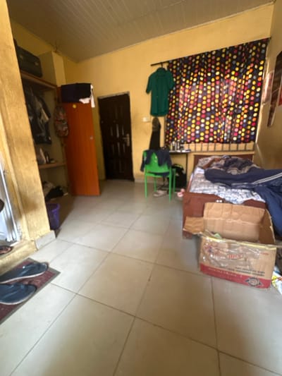 The image displays a moderately cluttered residential room located in Ado-Ekiti, Nigeria. The room features light yellow-painted walls and a ceiling composed of white slatted panels. The floor is covered with light-colored square tiles.

On the right side of the room, a bed with a dark brown wooden frame is positioned against the wall. It is covered with a white and blue patterned sheet, overlaid with dark blue blankets or folded clothing. Adjacent to the bed, a small black table holds several indistinct items. A large, open cardboard box, partially crushed and displaying red and white branding, rests on the tiled floor next to the bed.

A window on the right wall is covered by a multi-colored curtain featuring a polka-dot pattern in red, yellow, orange, and blue. A green collared shirt is hanging on the wall above the curtain rod, and a black shoe is visible hanging near the curtain. A green plastic chair stands in the midground, draped with dark clothing, with a pair of white sneakers positioned beneath it.

On the back wall, a dark brown security door with a silver-colored handle and lock mechanism is centrally located. To its left, a tall, narrow reddish-brown wooden door or cupboard is visible. Further to the left, a built-in wooden wardrobe or shelving unit contains various pieces of clothing and a patterned backpack. A dark purple bucket or container is present on the floor in the lower-left corner near the wardrobe.

In the immediate foreground, two dark-colored flip-flops are placed on a red mat. No individuals are present in the scene, and no specific actions or interactions are depicted. The room appears lived-in.