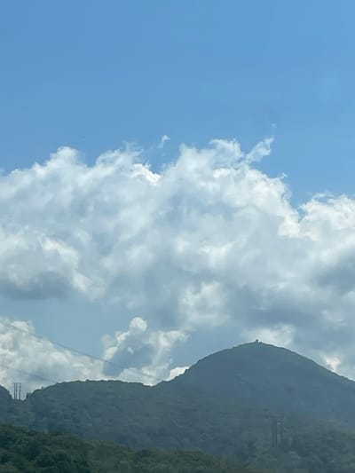 The image displays a hazy, mountainous landscape under a bright blue sky filled with large, puffy white clouds. In the foreground, lush green hills are covered in dense foliage. The middle ground features a prominent, rounded mountain peak with a small, dark structure, possibly a building or monument, situated at its summit. Another mountain range, also covered in trees, is visible to the left. Several tall, lattice-like transmission towers and power lines traverse the landscape, indicating the presence of infrastructure. The overall scene suggests an outdoor, natural environment, possibly in a rural or semi-rural area. The specific location in the city of Razdolnoye, Russia, is not visually identifiable from the provided image.