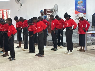 The image depicts an indoor scene in Goni Gara, Nigeria, featuring a group of approximately 12 young individuals, predominantly adolescents and young adults, alongside two visible adult women and one partially obscured adult man. The young participants are uniformly dressed in red long-sleeved shirts with black collars and cuffs, each bearing a white cross symbol on the chest, paired with black pants or skirts. Their footwear includes various sandals, slides, and a few pairs of white sneakers. Most of these individuals are positioned in a row, with their heads bowed slightly downwards and hands either on their hips or clasped in front, suggesting a synchronized posture for a performance or religious observance.
In the mid-ground, a woman wearing a blue and white patterned headwrap and dress stands behind a portion of the young group, observing them. To the far right of the frame, another adult woman, also in a red shirt with a white cross and a white headwrap, stands with her hands on her hips, facing forward. In the background, a large blue banner is prominently displayed, which reads "2026 Theme ENTERING INTO NEWNESS Through THE FRUIT AND GIFTS OF THE SPIRIT Acts 1:8," along with additional text in what appears to be a local language. A white wall-mounted oscillating fan is visible on the upper left wall, and another darker fan is positioned near a brown wooden door in the mid-background. A red-framed window with a metal security grille is present on the far left side of the room. The floor is covered with light-colored terrazzo or tiles, and the walls are plain white. A raised platform with grey steps is visible on the right side of the background, indicating a stage or altar area. A black furniture armrest and a small white side table are visible on the far right.