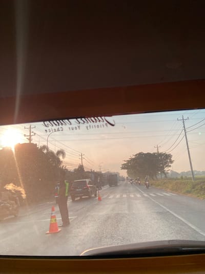 This image captures a road scene at sunrise in Jekulo, Indonesia. A person in a high-visibility vest stands near traffic cones on the right side of a two-lane road. Several vehicles, including cars and motorcycles, are visible on the road, with some appearing to be in traffic. The sun is low in the sky, casting a warm glow and creating lens flare. Power lines and trees line the sides of the road, and a pedestrian crossing is marked on the pavement. A blurry inscription is visible on the interior of the vehicle from which the photo was taken.