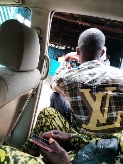 The image, captured from inside a vehicle in Ore, Nigeria, depicts an outdoor scene with multiple individuals. In the foreground, hands wearing yellow and black patterned attire are seen holding a black smartphone with a red case. A light-colored car seat headrest and a car door pillar displaying an "SRS CURTAIN AIRBAG" label are visible on the left. The central subject, viewed from behind, is a person with short grey hair, wearing a brown plaid shirt featuring a large golden "LV" logo on the back and a lighter patterned yoke. They are seated or standing, facing away from the camera, and wearing dark trousers. In the background, under a corrugated metal roof, an open-sided structure with blue wooden slat walls houses several people. A bright blue plastic bucket is prominent on the left, and various small bottles are visible on a surface. The ground appears paved. No direct interaction between the foreground individual and the central subject is apparent.