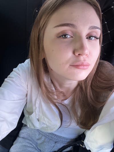 A close-up, high-angle selfie of a young Caucasian female with light brown hair and light green eyes. She is looking directly at the camera. She is wearing a white hooded jacket over a light gray t-shirt and gray sweatpants. Her hair is long and wavy, framing her face. She is wearing subtle makeup, including mascara and lip gloss. The background is dark and indistinct, suggesting an indoor setting, possibly a car interior, with some quilted texture visible on the right. There are no visible landmarks or environmental cues to indicate the specific location within the city of Tolbazy, Russia.