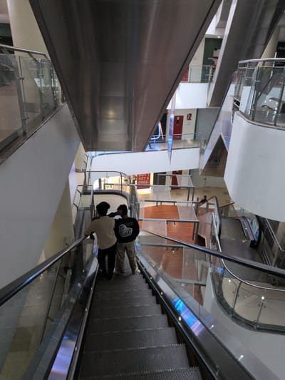 The image depicts the interior of a multi-story commercial complex or mall located in Hyderabad, India. The perspective is from an upper level, looking down across multiple floors connected by a series of escalators.

In the immediate foreground, two males stand on a descending escalator. The individual on the left wears a light-colored, long-sleeved shirt and dark trousers, while the individual on the right wears a dark, long-sleeved shirt with a graphic design on the back and light-colored trousers. Both are positioned with their backs to the viewer, observing the lower levels of the building. The escalator features dark, ribbed steps, a black handrail, and transparent glass side panels reflecting internal blue and purple light.

The midground and background reveal a spacious, modern architectural environment. Multiple escalators, both ascending and descending, traverse the different floors. Each level is bordered by contemporary glass and metal railings. Walls are predominantly white or light grey, with sections featuring distinct patterns, such as a geometric motif on a wall to the mid-right, and an accent wall of dark green or grey to the upper left. A prominent sign on a lower level advertises "BUY 1 GET 2 FREE," indicative of a retail establishment. Several red doors, potentially exits or service entrances, are visible on various floors. Large, angled metal structures cut diagonally through the space, supporting the levels or serving as decorative elements. A third individual is faintly visible on a lower level towards the right, near another escalator. The entire complex is illuminated by artificial lighting, creating a bright and open atmosphere.