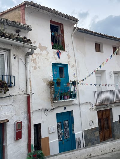 The image depicts a narrow street scene in Buñol, Spain, featuring three multi-story buildings under an overcast sky.
The central building, bearing the number '8' below its middle-floor balcony, has a weathered white facade with peeling paint and a terracotta tiled roof. The top floor features an open window with a red frame and a window box containing green foliage and several colorful pinwheel decorations. Directly below this window, a small red and blue diamond-shaped kite is affixed to the wall. The middle floor has a teal-blue double door with decorative grille work, opening onto a small balcony with a black metal railing. This balcony holds numerous potted plants, some with pink blooms, and two white pinwheel decorations. A white vertical drainpipe runs down the left side of this building, turning red at its lower section. The ground floor presents another teal-blue double door with similar decorative grille work. To the left of this door, a light-colored wooden board leans against the wall, and a red electrical box is mounted further left.
To the right, a multi-story building marked with the number '6' on its ground floor. Its upper section has a white facade, transitioning to dark grey, slate-like tiles on the lower half. The top floor features two small, dark-framed windows. The middle floor has two windows with closed white shutters and a balcony with a black metal railing, devoid of plants. The ground floor has a brown wooden double door with a small grille. A string of colorful, crescent-shaped fabric bunting hangs diagonally across the street, connecting the central building to this building on the right.
To the far left, a partially visible white building features a window with a blue metal balcony containing potted plants. No people are visible in the scene. The street surface is partially visible at the bottom, suggesting a curb or low wall in the foreground.