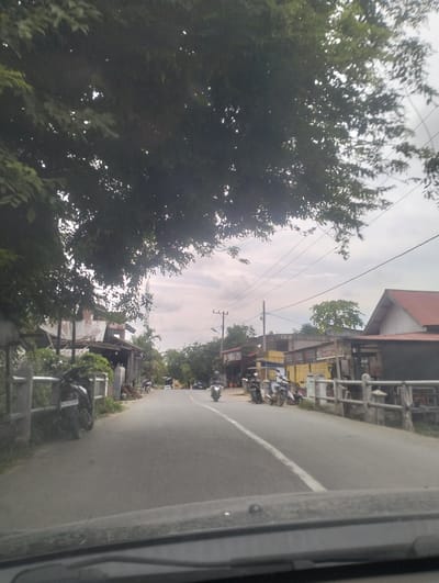 The image depicts a street scene in Muliengmanyang, Indonesia, taken from a vehicle. The foreground shows the dashboard and windshield of the vehicle. The road ahead is paved with asphalt and has a white line marking the center. On the left side of the road, there is a gray metal fence and some vegetation. Beyond the fence, there are buildings with corrugated metal roofs, one of which has a satellite dish. Several motorcycles are parked on the side of the road, and one is in motion further down the street. On the right side of the road, more buildings with red and orange roofs are visible. Overhead power lines crisscross the sky, which is overcast with clouds. Trees with dense foliage are present throughout the scene, casting shadows on the road and buildings. The overall impression is of a moderately built-up area on a cloudy day.