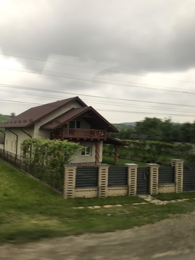 The image depicts a residential scene in Cristești, Romania, under an overcast sky. A two-story house with light-colored walls and a multi-gabled dark brown tiled roof is the central subject. The house features a second-story balcony with a wooden railing and supports, positioned above a smaller covered porch area. A green hedge or row of bushes is visible in front of the house's lower section. In the immediate foreground, a fence composed of dark, possibly corrugated metal panels and light-colored brick pillars runs horizontally, separating the property from a dirt or gravel path. Green grass slopes upward from the path towards the house. The background includes more green foliage and distant hills under a uniformly grey, cloudy sky. Several horizontal utility lines are visible in the upper portion of the frame. No people or animals are present.